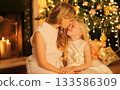 Happy mother kissing her child daughter near Christmas tree and fireplace at home 133586309
