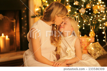 Happy mother kissing her child daughter near Christmas tree and fireplace at home Happy mother kissing her child daughter near Christmas tree and fireplace at home 133586309