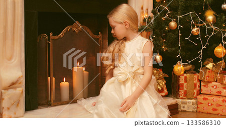 Happy little girl child with festive gift box sitting near Christmas tree and fireplace at home Happy little girl child with festive gift box sitting near Christmas tree and fireplace at home 133586310