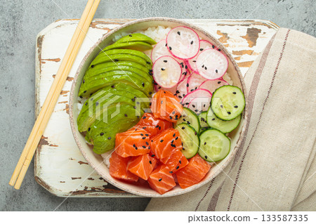 Salmon poke bowl with avocado, radish, cucumber and rice on white rustic wooden board on grey background top view 133587335