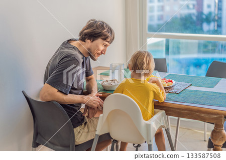 A father having breakfast at the table with his young son in a bright home kitchen, sharing a warm morning moment. Family bonding, parenting, everyday routine and healthy lifestyle concept 133587508