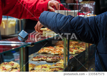 A customer receives a fresh baked flatbread snack from a vendor in red uniform behind a glass counter filled with assorted warm festive street food at a winter market. 133587573