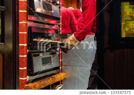 Street market scene showing baker pulling a tray of food from an industrial oven at a food market or bakery stand. 133587575