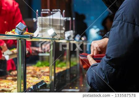 Customer paying for hot street food opening a wallet while a vendor in a red jacket serves hot snacks behind a glass counter at a winter market stall Customer paying for hot street food opening a wallet while a vendor in a red jacket serves hot snacks behind a glass counter at a winter market stall 133587578