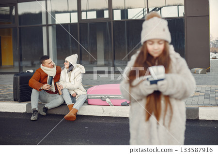 Little girl with her parents going on vacation. Portrait of a girl standing in front of photo and and her parents sitting behind. Family wearing warm clothes. 133587916