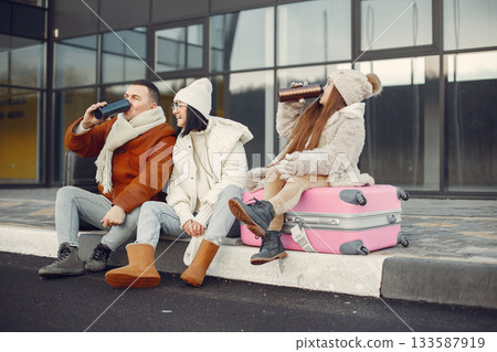 Family of three going on vacation. Man and girl sitting on a luggage and drinking tea from thermos. Mother, father and daughter wearing warm clothes. Family of three going on vacation. Man and girl sitting on a luggage and drinking tea from thermos. Mother, father and daughter wearing warm clothes. 133587919