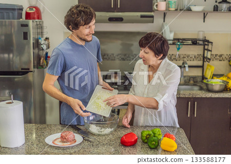 Man preparing lunch together with his elderly mother in an authentic modern kitchen, cutting ingredients and cooking a homemade meal in a warm family atmosphere, concept of family care and quality 133588177