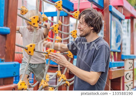 A man playing with his small son on a playground, laughing and engaging in active fun together in a bright outdoor setting. Family bonding, early childhood development and healthy outdoor lifestyle 133588190