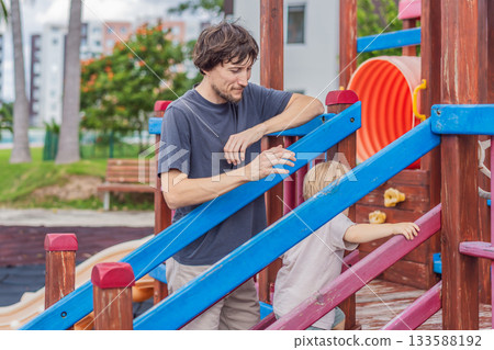 A man playing with his small son on a playground, laughing and engaging in active fun together in a bright outdoor setting. Family bonding, early childhood development and healthy outdoor lifestyle 133588192