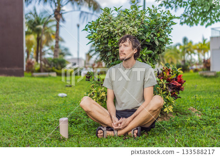 A man relaxing in a park on a sunny day, sitting peacefully on the grass and enjoying nature in a calm outdoor setting. Leisure time, mindfulness, mental wellness and slow living lifestyle concept 133588217
