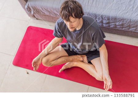 Man practicing yoga at home on a mat, performing calm stretching exercises in a cozy living room, concept of home fitness, mindfulness, balance, health and emotional well-being Man practicing yoga at home on a mat, performing calm stretching exercises in a cozy living room, concept of home fitness, mindfulness, balance, health and emotional well-being 133588791