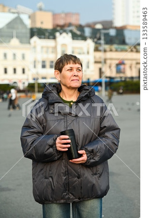 60 year old russian woman with short hair, holding thermos cup with coffee standing in city Vladivostok and lookind right 133589463