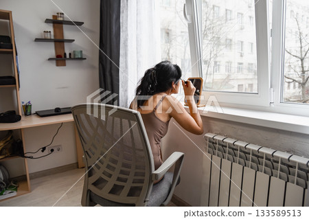 Russian 50 year old woman seen from behind sitting in a bedroom holding a brow brush preparing her eyebrows in a quiet calm moment of personal beauty care 133589513