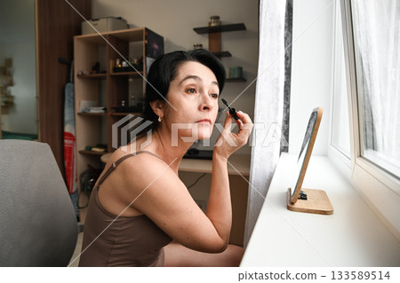 Close view of a Russian 50 year old woman applying mascara showing natural wrinkles and focused expression during her quiet beauty routine at home 133589514