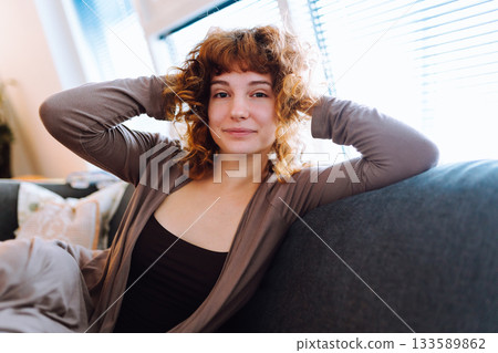 Smiling positive young woman sitting on sofa near window at home Smiling positive young woman sitting on sofa near window at home 133589862