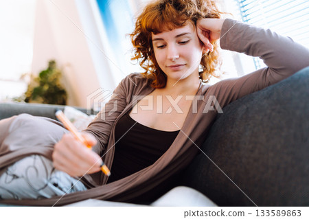young woman at home writing notes in notebook while sitting on sofa young woman at home writing notes in notebook while sitting on sofa 133589863
