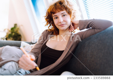 young woman at home writing notes in notebook while sitting on sofa young woman at home writing notes in notebook while sitting on sofa 133589868