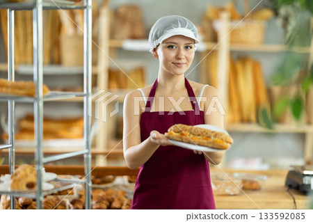 Happy young pretty girl baker in apron with joyful expression holds freshly baked bocadillo in bakery shop, showcasing concept of food industry and catering services 133592085