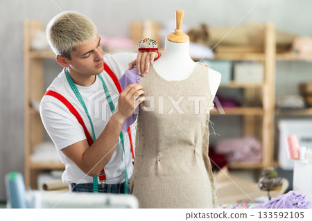 Experienced young male fashion designer working with fabric and mannequin while sewing fashionable dress in sewing workshop Experienced young male fashion designer working with fabric and mannequin while sewing fashionable dress in sewing workshop 133592105