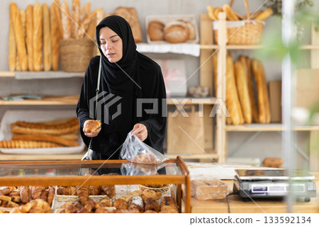 Girl in traditional Muslim clothing selects and places fresh hot croissants in plastic bag 133592134