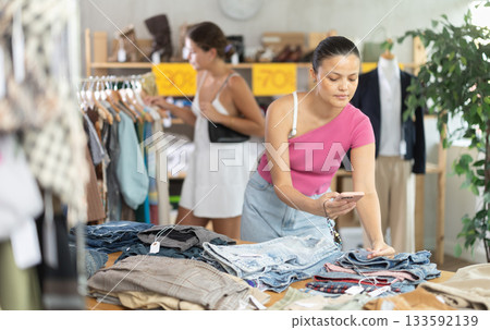 Attentive young Asian woman scanning QR-code on pantaloons in clothing store 133592139