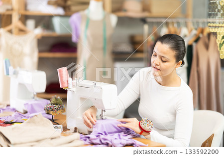 Professional young attentive woman seamstress sewing textile using sewing machine sitting at table at workplace in sewing studio 133592200