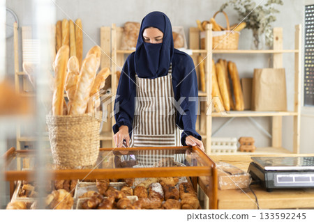 Muslim girl seller employee puts croissants in window, arranges display of goods at bakery. 133592245