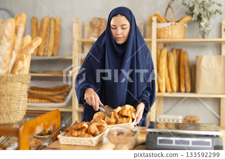 Muslim girl seller employee puts croissants in window, arranges display of goods at bakery. Muslim girl seller employee puts croissants in window, arranges display of goods at bakery. 133592299