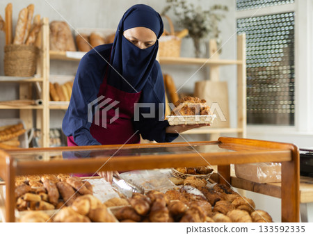 Asian Muslim girl seller employee puts croissants in window, arranges display of goods at bakery. Asian Muslim girl seller employee puts croissants in window, arranges display of goods at bakery. 133592335