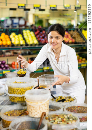 Satisfied young woman putting pickled olives into a container in the grocery section of supermarket Satisfied young woman putting pickled olives into a container in the grocery section of supermarket 133592359