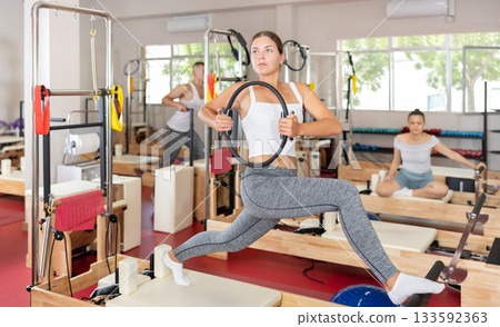 Sportive young girl training her arms and legs on reformer device Sportive young girl training her arms and legs on reformer device 133592363