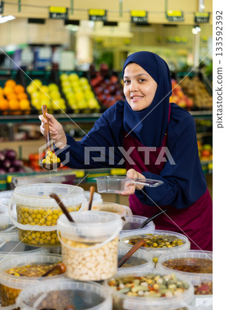 Female seller in hijab and an apron packs pickled olives in container in the grocery section of supermarket Female seller in hijab and an apron packs pickled olives in container in the grocery section of supermarket 133592392