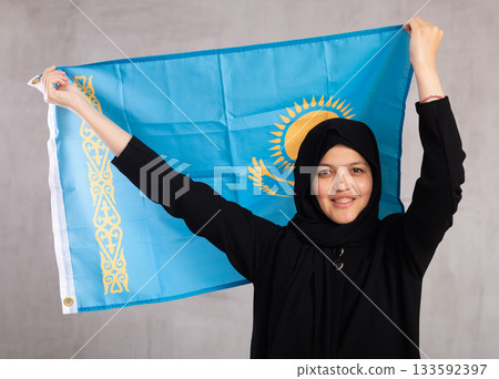 Young Muslim woman in black hijab holds unfurled flag of Kazakhstan 133592397