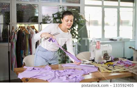 Positive young Asian tailoress cutting textile in sewing studio Positive young Asian tailoress cutting textile in sewing studio 133592405