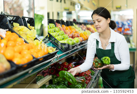 Positive asian girl seller in an apron offers green bell peppers Positive asian girl seller in an apron offers green bell peppers 133592453
