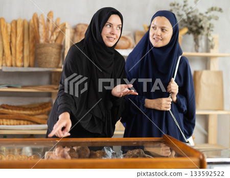 Two girls in traditional Muslim clothing chat and choose delicious bread and croissants together at bakery Two girls in traditional Muslim clothing chat and choose delicious bread and croissants together at bakery 133592522