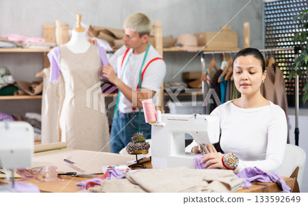 Attentive woman tailor sewing textile using sewing machine and young male assistant working with fabric and mannequin in fashionable sewing atelier Attentive woman tailor sewing textile using sewing machine and young male assistant working with fabric and mannequin in fashionable sewing atelier 133592649