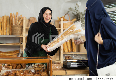 Selling fresh bread in an oriental bakery - girl seller hands bag of baguettes to female customer Selling fresh bread in an oriental bakery - girl seller hands bag of baguettes to female customer 133592656