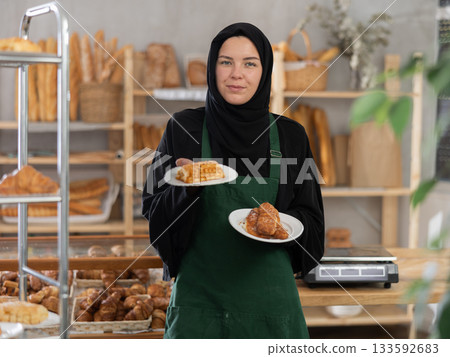 Positive girl seller of oriental bakery in traditional Muslim clothes with fresh hot pastries in hands Positive girl seller of oriental bakery in traditional Muslim clothes with fresh hot pastries in hands 133592683