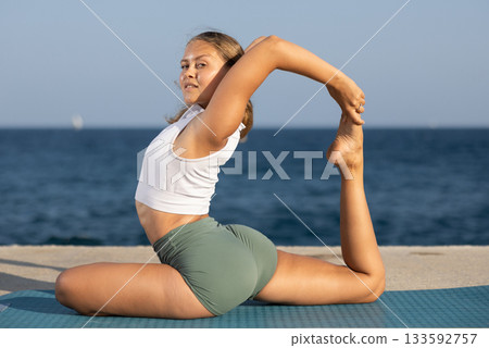 Sportive girl practicing king pigeon pose of yoga on seashore 133592757