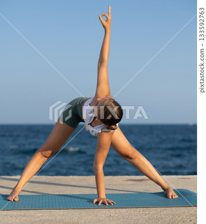Sportive girl practicing revolved forward bend pose of yoga on seashore 133592763