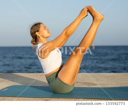 Sportive girl practicing boat pose of yoga on seashore Sportive girl practicing boat pose of yoga on seashore 133592773