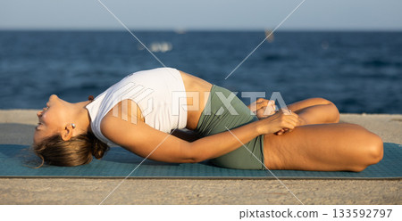 On windy day young slender woman does yoga near sea doing matsiasana 133592797