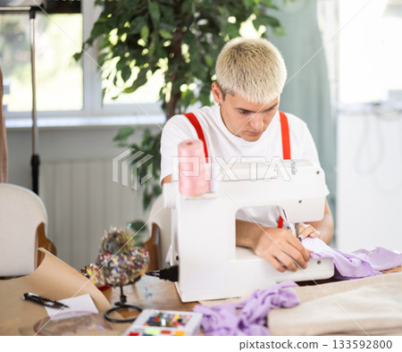 Attentive man tailor sewing textile using sewing machine sitting at workplace in fashionable sewing atelier 133592800