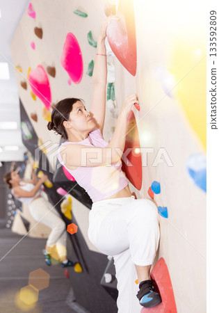 Asian girl is focused on climbing to top of artificial bouldering wall along complicated route track Asian girl is focused on climbing to top of artificial bouldering wall along complicated route track 133592809