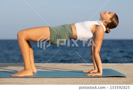 On windy day young slender woman does yoga near sea doing reverse plank 133592825