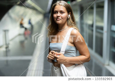 Girl with earphones is standing on subway platform, waiting for train to arrive 133592903