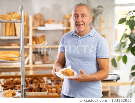 Senior male customer stands with croissant in hands near window of bakery. Senior male customer stands with croissant in hands near window of bakery. 133592935