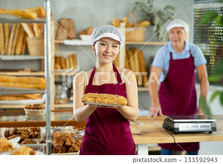 Positive young girl baker in white uniform baking and offering to buy fresh bocadillo in bakery, elderly male baker working in background 133592943