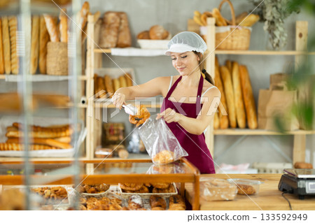 Young girl seller in apron putting fresh croissant in plastic bag in bakery shop 133592949